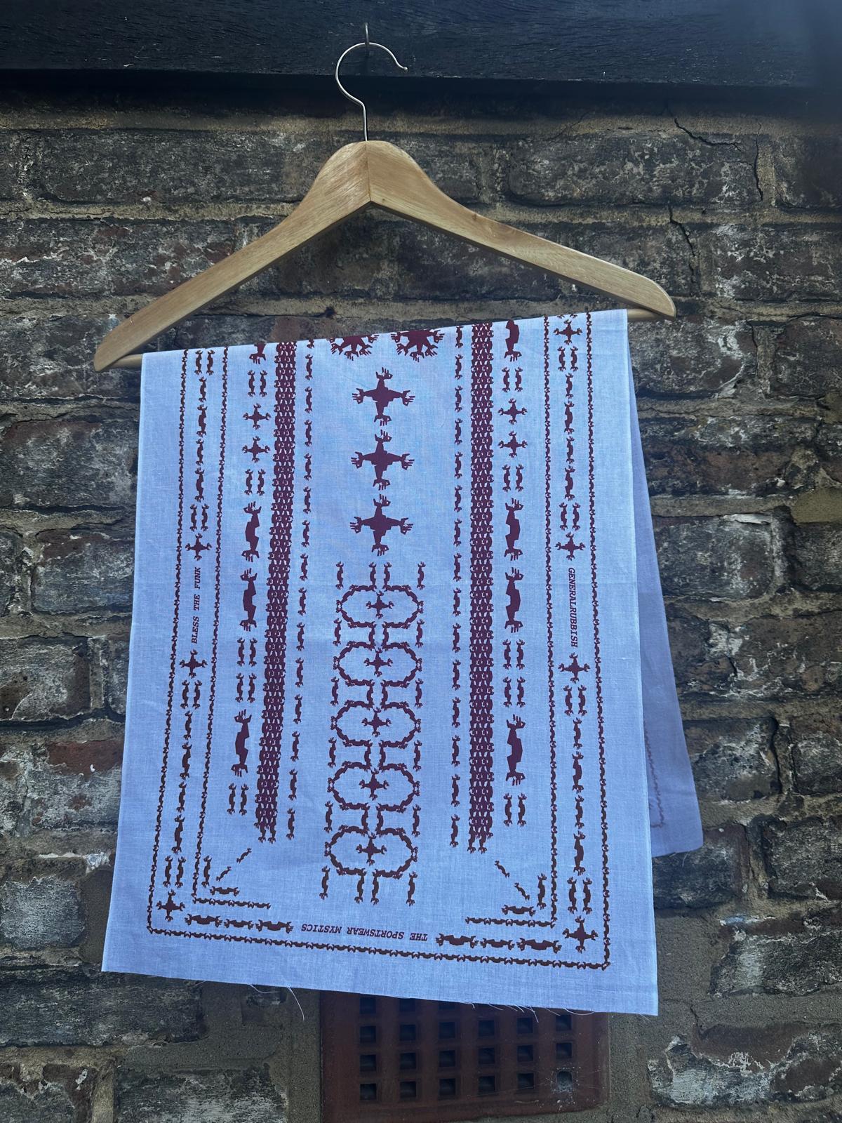 A white tenugui with goji berry red screen printed Galliano hand patterns hanging from a wooden hanger against a urban brick wall.