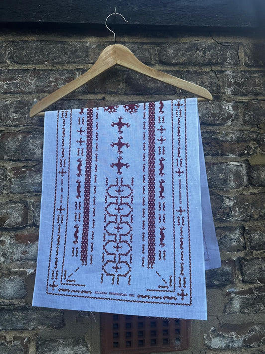 A white tenugui with goji berry red screen printed Galliano hand patterns hanging from a wooden hanger against a urban brick wall.